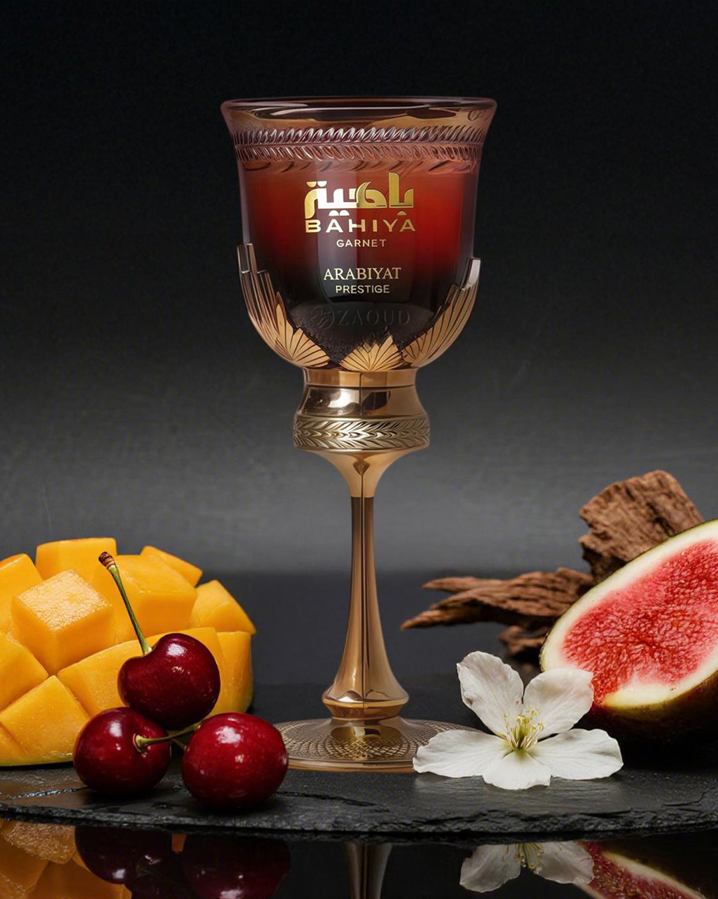 A deep-red perfume bottle of Bahiya Garnet by Arabiyat Prestige, stands elegantly on a black surface. Around it is diced mango, ripe cherries, half a fig, soft white blossom, and aromatic wood.