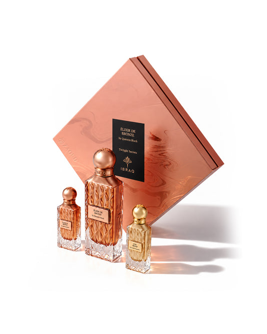 Ibraq elixir de bronze twilight secret set with dor tresor perfume bottles with metallic rose-gold and gold caps arranged in front of a matching rose-gold gift box on a clean white background. The central bottle is larger and textured, with two smaller matching bottles on either side.
