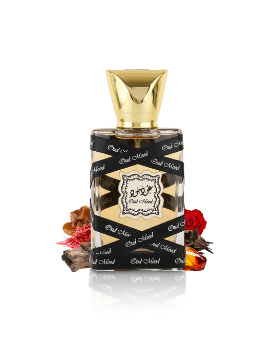 lattafa oud mood miniature perfume bottle with a faceted gold cap, wrapped in a black ribbon printed with 'Out of Now' in white script. The bottle is styled with dried botanicals and red rose petals on a reflective white surface.