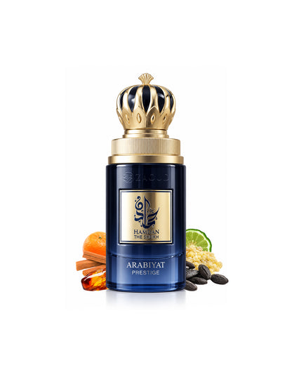 A bottle of Arabiyat prestige hamdan the sheikh, featuring a deep blue bottle with a gold crown cap, surrounded by its scent ingredients including orange, cinnamon, black seed, and lime on a white background.