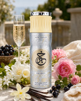 A bottle of Khair Felicity perfume spray is centered on a styled outdoor table setting with a champagne flute, flowers, vanilla beans, and dark berries. The product bottle is metallic silver with a cream-colored cap and a gold circular logo.