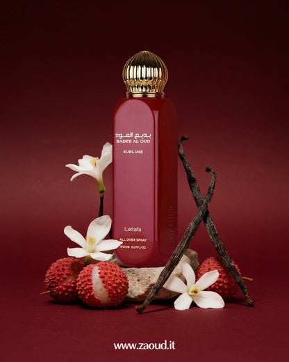 Badee Al Oud Sublime by Lattafa, placed with white flowers, lychee fruits, vanilla beans, and a stone, against a dark red background.