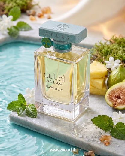 A bottle of Lattafa Atlas Glacial Valley perfume is displayed on a marble slab in a turquoise pool, surrounded by mint leaves, white flowers, a pineapple slice, and a fig.