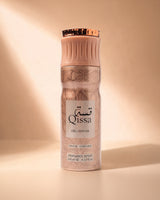 A perfume spray bottle from Qissa Delicious, Paris Corner, standing upright on a matching surface. The bottle has a warm beige and rose gold metallic pattern with a white label and a metallic rose gold cap, set against a warm beige background with soft diagonal light rays.