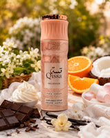 A bottle of Qissa Delicious perfume spray stands centered on a styled surface with chocolate bars, whipped cream, marshmallows, vanilla beans, flowers, and citrus slices. The bottle is pale pink with a metallic rose-gold cap and a cream label.