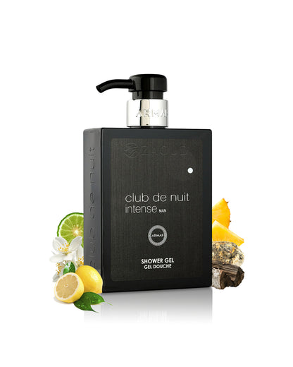 A black rectangular shower gel bottle with a black pump dispenser, labeled 'club de nuit intense MAN', styled with citrus fruits, a lime slice, white flowers, and bark on a white background.