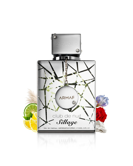 Armaf Club de Nuit sillage Eau de Parfum bottle with a silver cap and a geometric black and green patterned glass body, styled with citrus slices, flowers, and pampas grass on a white background.