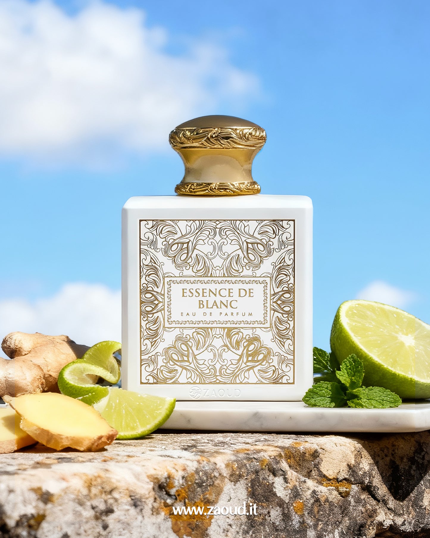 Close-up of the luxurious 'Essence de Blanc' Eau de Parfum bottle by fragrance world. The square white bottle features intricate gold filigree and an ornate gold cap, and is set outdoors on a stone surface under a bright blue sky. Fresh ingredients like slices of lime, ginger root, and sprigs of mint leaves are arranged around the base, suggesting the fragrance notes.