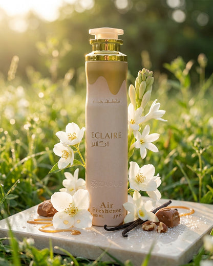 lattafa Eclaire Air freshener is styled outdoors on a stone slab with white flowers, vanilla beans, and nuts, set against a blurred green grassy background with warm sunlight.