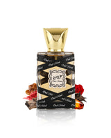 lattafa oud mood miniature perfume bottle with a faceted gold cap, wrapped in a black ribbon printed with 'Out of Now' in white script. The bottle is styled with dried botanicals and red rose petals on a reflective white surface.