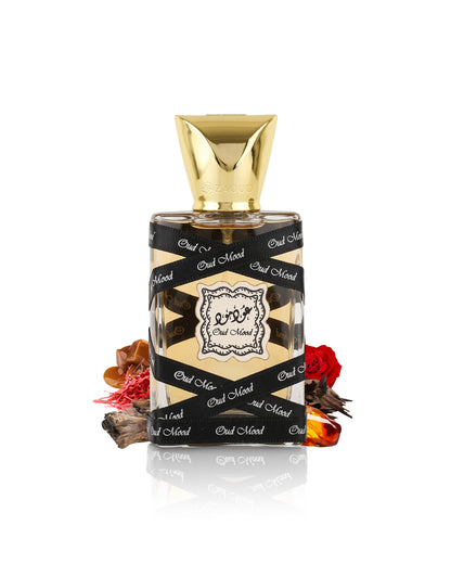 lattafa oud mood miniature perfume bottle with a faceted gold cap, wrapped in a black ribbon printed with 'Out of Now' in white script. The bottle is styled with dried botanicals and red rose petals on a reflective white surface.
