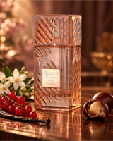 A faceted clear glass perfume bottle with a warm pinkish-amber tint and a central label reading 'KHAMRAH mira lattafa', styled on a reflective wooden surface with red berries, white flowers, vanilla bean, and chestnuts in a softly lit setting.