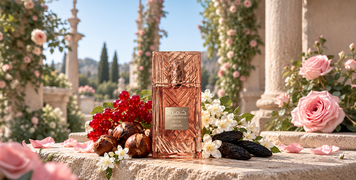 A luxury perfume bottle with a textured metallic rose-gold/copper finish stands on a stone terrace in a garden setting, surrounded by pink roses, red berries, and dark olives. The background features classical columns and arches under a clear blue sky.