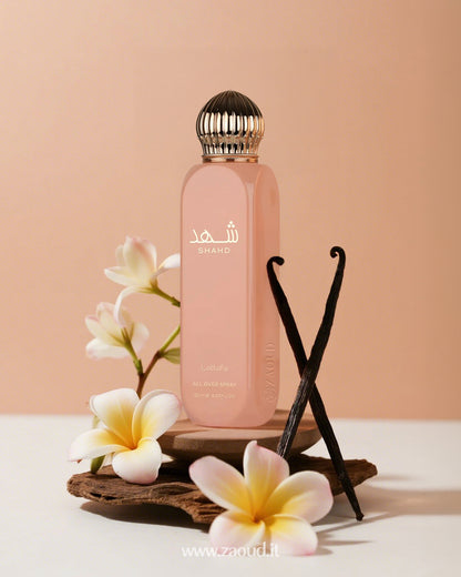Lattafa Shahd All Over Spray, placed on a wooden surface with vanilla sticks and white flowers around it. The background is a soft peach color.