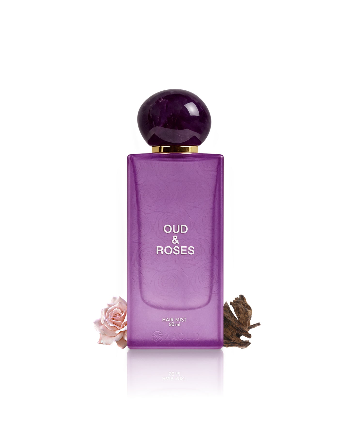 A purple rectangular bottle of Oud & Roses Hair Mist 100ml with a dark purple spherical cap and gold collar, displayed on a white background with decorative dried rose and pink rose elements at the base.