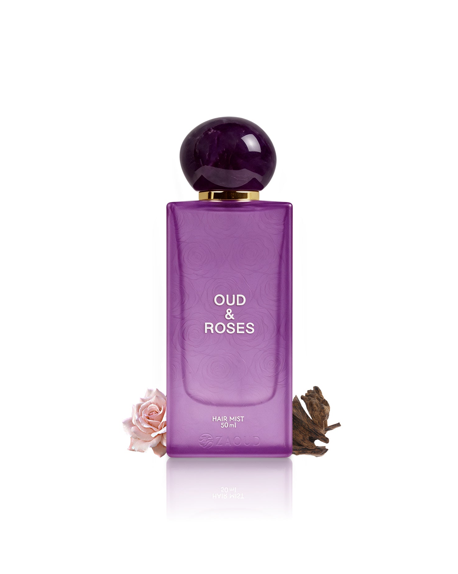 A purple rectangular bottle of Oud & Roses Hair Mist 100ml with a dark purple spherical cap and gold collar, displayed on a white background with decorative dried rose and pink rose elements at the base.