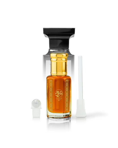 A clear glass tambul perfume oil bottle with amber-orange liquid, a black cap, and a gold-toned neck, shown on a white background with a small stick applicator wand beside it.